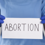 Abortion - California Family Council Person wearing blue gloves and scrubs holds a white sign with the word 'ABORTION' written in large letters.