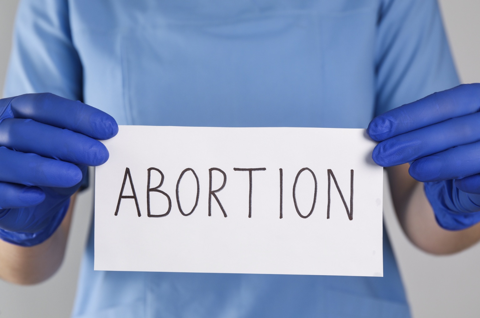 Abortion - California Family Council - California Family Council Person wearing blue gloves and scrubs holds a white sign with the word 'ABORTION' written in large letters.