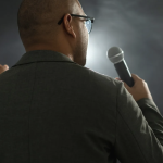 Back view of a man in a suit holding a microphone, gesturing with his other hand on a dim stage backdrop.