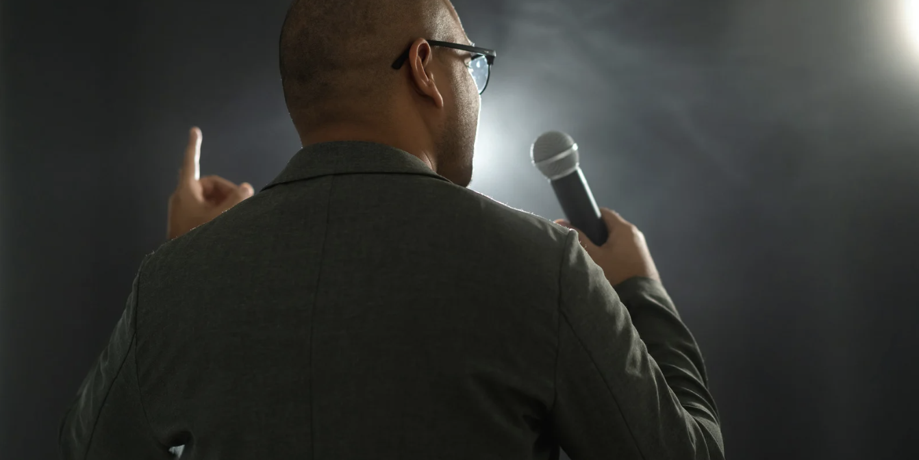 speaker - California Family Council - California Family Council Back view of a man in a suit holding a microphone, gesturing with his other hand on a dim stage backdrop.