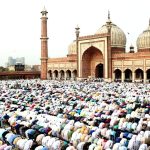 Massive crowd of worshippers praying in the courtyard of a grand mosque with domes and minarets, many wearing white robes and colorful outfits.