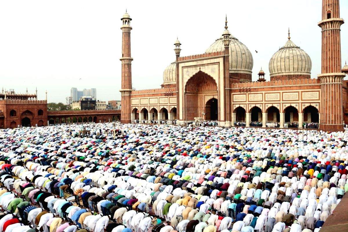 Massive crowd of worshippers praying in the courtyard of a grand mosque with domes and minarets, many wearing white robes and colorful outfits.