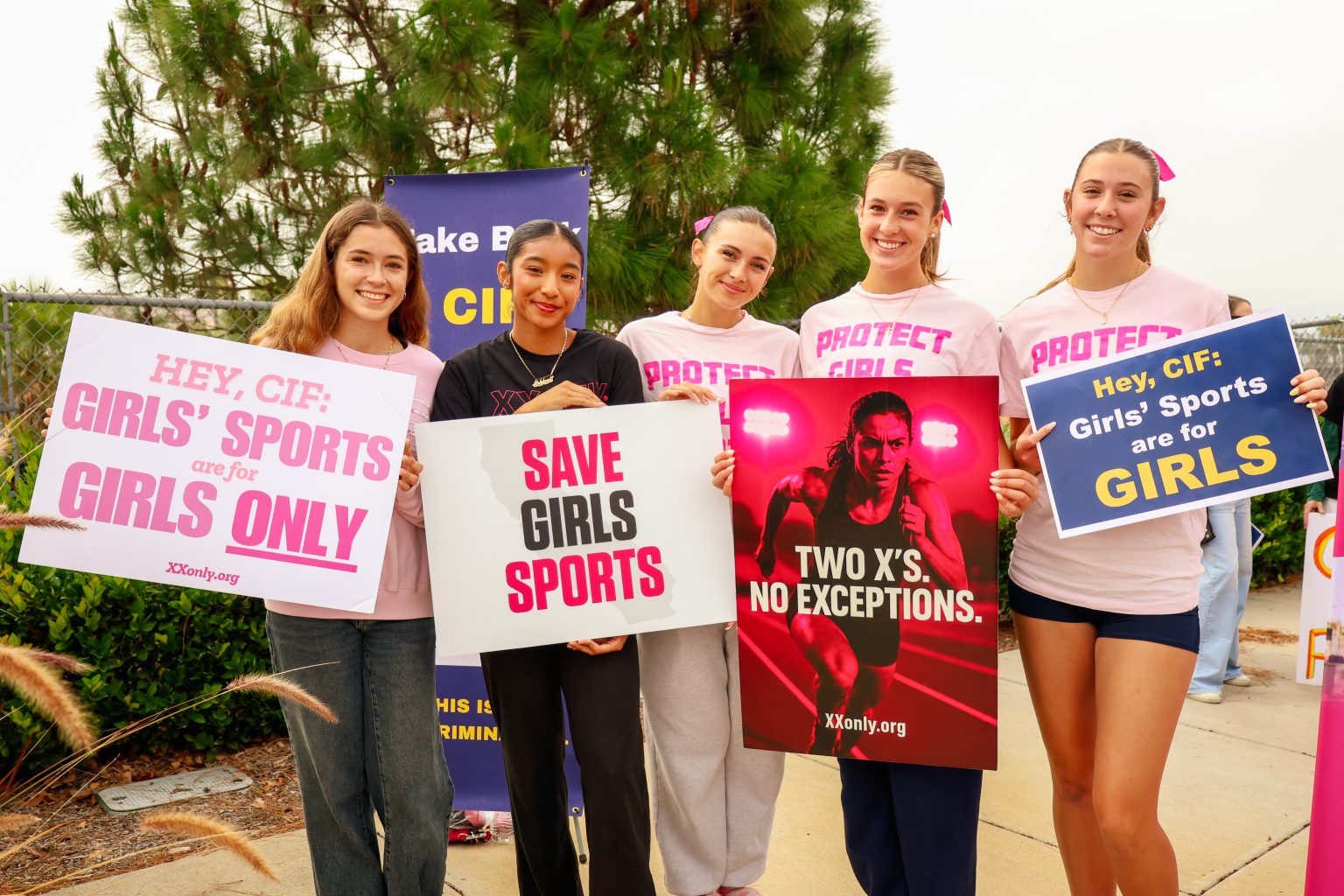 Group of young women holding signs supporting girls' sports at a rally, smiling at the camera.
