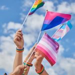 Several hands with rainbow bracelets raise LGBTQ+ pride flags against a blue sky.