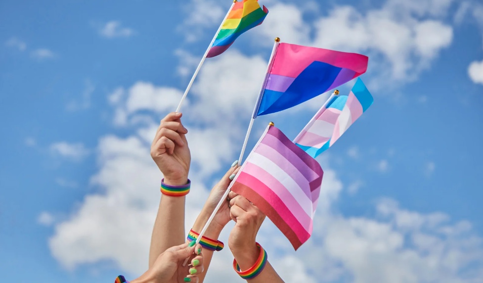 Several hands with rainbow bracelets raise LGBTQ+ pride flags against a blue sky.