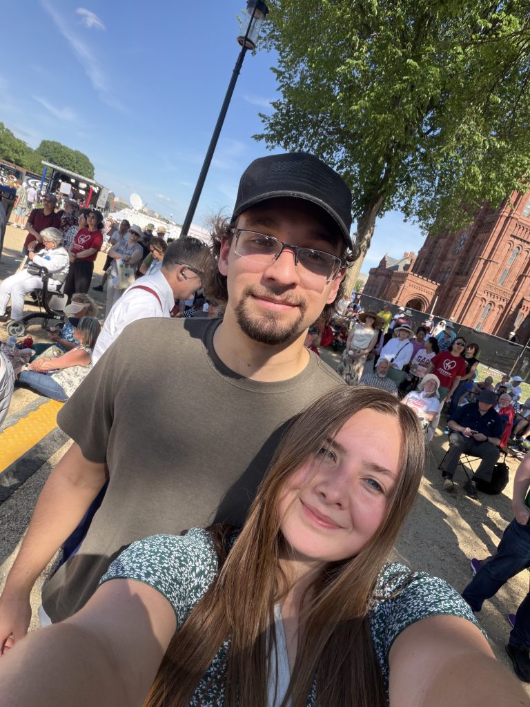 Two people take a sunny selfie at a crowded outdoor event, with a brick building and trees in the background.