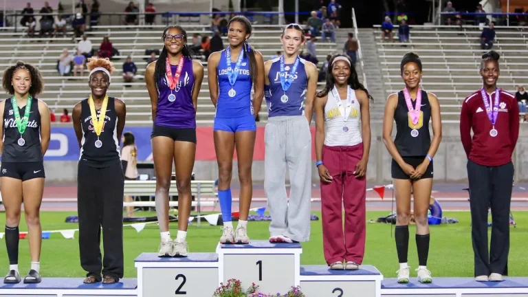 Group of young female athletes on a podium after a track event, wearing medals and posing for photos.