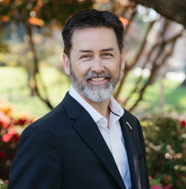 Smiling man in a dark blazer and white shirt posing outdoors in a park with trees in the background, sunlight filtering through leaves.