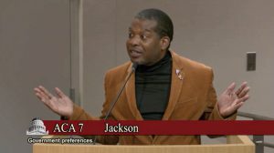Man in a brown blazer speaks at a podium with hands raised; banner reads 'ACA 7 Jackson' and 'Government preferences' in foreground.