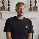 Young man with blond hair in a black Carhartt t-shirt sits in a living room in front of a white fireplace, with plants nearby.