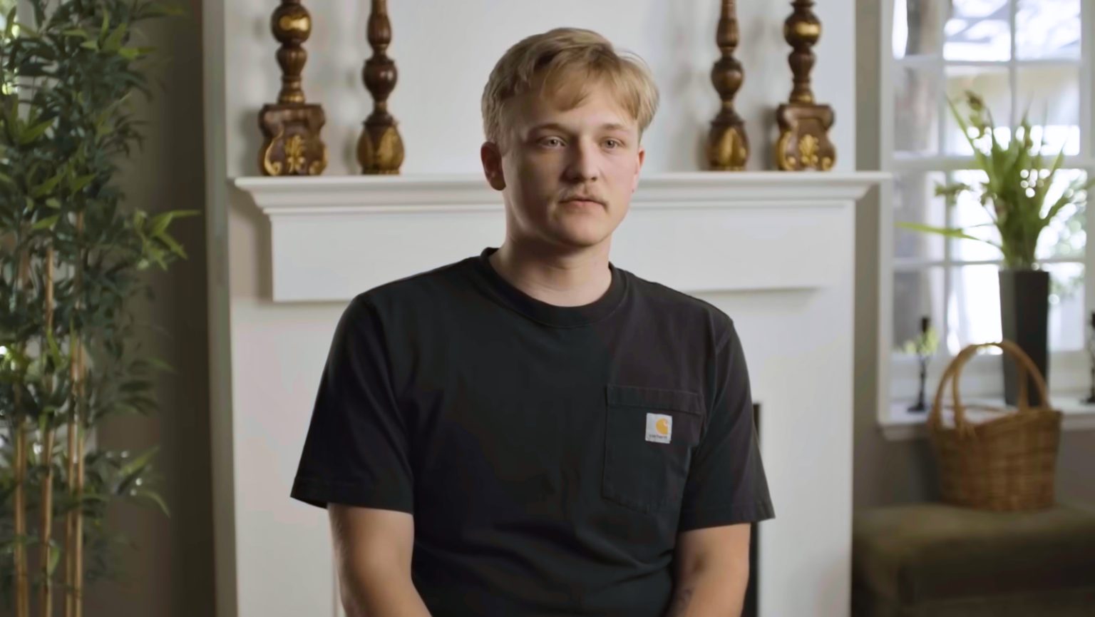 Young man with blond hair in a black Carhartt t-shirt sits in a living room in front of a white fireplace, with plants nearby.