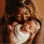 Mother with curly hair gently cradling a peacefully sleeping newborn in a white swaddle against a warm brown background.