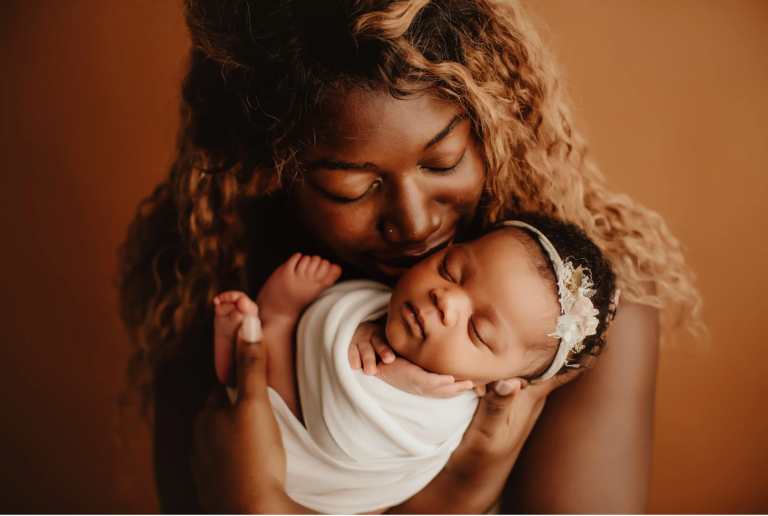 Mother with curly hair gently cradling a peacefully sleeping newborn in a white swaddle against a warm brown background.