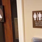 All gender bathroom - California Family Council Brown wall sign reads 'ALL-GENDER MULTI-USER RESTROOM' with male, female, and wheelchair icons beside a door area, indicating accessible facilities at the location. Left side shows a decorative square plaque with a white triangle inside a circle on the door.