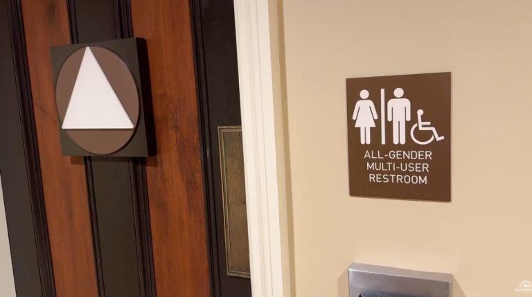 All gender bathroom - California Family Council - California Family Council Brown wall sign reads 'ALL-GENDER MULTI-USER RESTROOM' with male, female, and wheelchair icons beside a door area, indicating accessible facilities at the location. Left side shows a decorative square plaque with a white triangle inside a circle on the door.