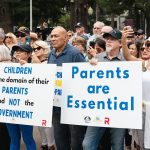 Crowd of protesters holding signs: 'Children are the domain of their parents and not the government' and 'Parents are Essential' at an outdoor rally, diverse participants visible with signs and phones raised.