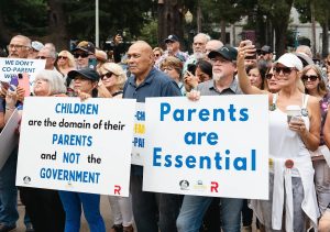 Crowd of protesters holding signs: 'Children are the domain of their parents and not the government' and 'Parents are Essential' at an outdoor rally, diverse participants visible with signs and phones raised.