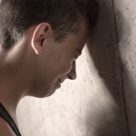 Young man with his head resting against a textured wall, eyes closed in a moment of reflection or sadness.