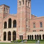 Brick university-style building with two tall towers, arched entry, and colonnade on a sunny day; people walking on the lawn nearby.