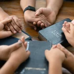 parying kids - California Family Council Several people hold hands in a circle around a Bible on a wooden table, symbolizing unity or prayer.